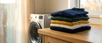 Stack of neatly folded colorful clothes on wooden surface near washing machine in bright laundry room for household chores and domestic cleaning tasks.