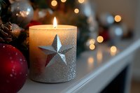 Glowing white candle decorated with silver star against blurred Christmas lights background, red ornament and pinecone creating festive holiday atmosphere.