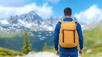 Male hiker with yellow backpack standing on mountain trail overlooking snow-capped peaks and green valley, wearing blue jacket on summer hiking adventure.