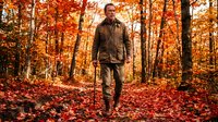 Man jogging through autumn forest with vibrant orange and red foliage, wearing brown athletic jacket and pants, surrounded by fallen leaves on ground.