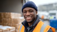 Young African American male warehouse worker wearing safety vest and winter hat smiling outdoors near shipping containers, expressing positivity at work.