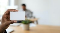 Male professional with dark skin tone holding blank white business card in office environment with blurred colleague and desk in background.