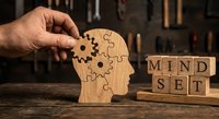 Male hand placing wooden puzzle piece into human head silhouette with gear mechanism next to mindset letter blocks on rustic wooden surface.
