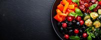 Fresh colorful vegetables and berries on black plate with basil leaves, cranberries, bell peppers and marinated olives against dark stone background.