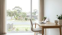 Minimalist home office workspace with wooden desk and beige chair by large window overlooking green suburban neighborhood for remote work productivity.