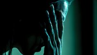 Dramatic silhouette of person covering face with hand in turquoise neon lighting, expressing emotional distress and vulnerability against dark background.