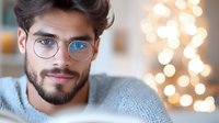 Young Caucasian man with beard and glasses smiling at camera, wearing cozy gray sweater against blurred bokeh lights background. Warm atmospheric portrait.