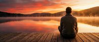 Man sitting on wooden dock at sunrise, watching misty lake with golden sky reflection, peaceful morning meditation and contemplation scene.