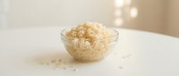 Cooked rice in glass bowl on white surface with scattered grains, suitable for food blogs, cooking websites, and culinary publications.