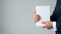 Businessman in dark suit holding blank white paper sheet against neutral gray background for corporate messaging and advertising purposes.