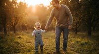 Father and toddler son walking hand in hand through sunny park during golden hour sunset creating warm family bonding moment outdoors in nature.