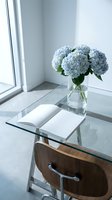 Minimalist home office with glass desk, open book, and blue hydrangeas in a vase by a bright window, modern interior design concept.