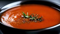 Creamy tomato soup garnished with fresh rosemary sprig and black pepper in dark ceramic bowl, close-up view showing smooth texture and vibrant red color.