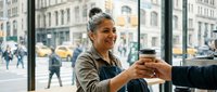 Middle-aged woman with gray hair in a bun, olive skin tone, wearing casual olive-green jacket, handing coffee cup to someone indoors near large window overlooking urban street with yellow taxis and pedestrians. Suitable for hospitality, service industry, lifestyle, and business communications.