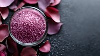 Pink crystal bath salt in glass jar with rose petals scattered on dark stone background, spa wellness concept for beauty and aromatherapy treatments.