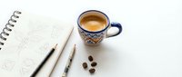 Overhead view of workspace with sketchbook, pencils, coffee cup and beans on white surface.
