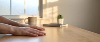 Hands resting on wooden table surface in bright modern interior with coffee cup and plant creating peaceful workspace atmosphere for relaxation.