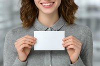 Smiling young Caucasian woman with wavy brown hair in grey business attire holding blank white card for your message in bright office environment.