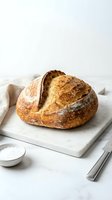 Freshly baked artisan sourdough loaf with golden crust on marble board, linen cloth, salt bowl and knife, rustic homemade bakery concept.