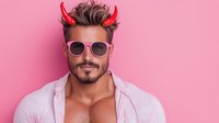 Young Hispanic man with devil horns and sunglasses smirking against pink background. Muscular male model wearing unbuttoned white shirt shows playful, flirtatious expression.