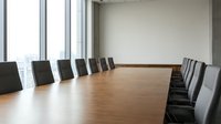 Modern corporate boardroom with long wooden conference table and black leather chairs overlooking city skyline through floor-to-ceiling windows.