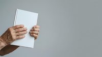 Hand holding blank white notebook or journal against neutral gray background, suitable for branding, mockups, and editorial designs.