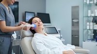 Professional cosmetologist performing facial treatment on relaxed female client in modern medical spa clinic with advanced equipment and sterile environment.