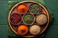 Colorful aromatic spices and seasonings in wooden bowls arranged on round wooden tray over green textile background, top view composition for cooking and food styling.
