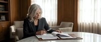 Senior woman with gray hair and light skin tone, wearing professional black blazer, reviewing documents at white desk in elegant office setting with wooden furniture and natural light from windows.
