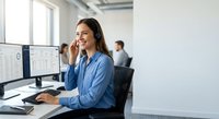 Young professional woman with headset working at computer in modern office environment, customer service representative smiling during call.