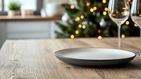 Empty gray ceramic plate on wooden table with blurred Christmas tree and wine glass in background, festive holiday dining setting with bokeh lights.