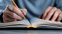 African American student writing in open notebook with yellow pencil, close-up view of hands on textbook pages showing focused study and learning process.