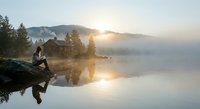 Young woman sitting on rocky shore at misty mountain lake during golden sunrise, peaceful morning reflection scene for wellness and nature concepts.