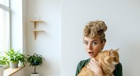 Woman with blonde hair styled in updo holding ginger cat indoors. Bright minimalist interior with potted plants and wooden shelves. Casual lifestyle portrait for home, pet, and design projects.