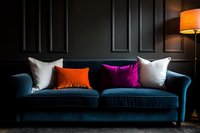 Luxurious teal velvet sofa with vibrant orange and purple accent pillows against dark paneled wall, illuminated by warm lamp light. Modern living room decor.