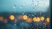 Raindrops on window glass with blurred city lights creating bokeh effect in blue and orange tones, moody atmospheric evening scene captured during rainfall.