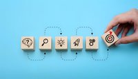 Hand placing wooden block with target icon completing a business strategy sequence on blue background, showing steps from idea to goal.