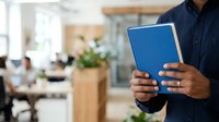 Man in dark shirt holding blue tablet in modern office workspace with colleagues and plants in background.