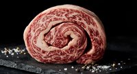Raw marbled beef roll on dark slate with coarse salt, premium wagyu meat cut showing intricate fat marbling patterns for gourmet cooking and culinary preparation.