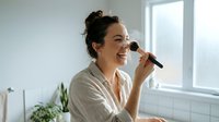 Young woman applying makeup with brush in bright bathroom, morning beauty routine, natural lighting, skincare and cosmetics lifestyle.