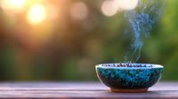 Aromatic smoke rising from turquoise ceramic bowl with burning incense on wooden surface against blurred natural background with sunset bokeh lights.