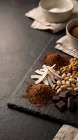 Cocoa powder, chocolate pieces, nuts and spices arranged on dark slate board with ceramic bowls in background for baking and confectionery preparation.