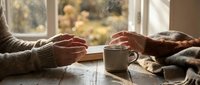Cozy winter morning scene with hands warming around hot coffee mug by window with Christmas tree lights creating peaceful holiday atmosphere at home.