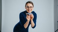 Young woman with glasses wearing dark blue blazer holding positive pregnancy test result, smiling at camera against white background.