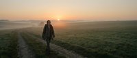 Person in dark jacket standing on rural dirt path at sunrise with misty fields and golden sky, suitable for travel, adventure, and lifestyle editorial use.