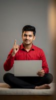 Young Indian businessman in red shirt pointing up with laptop computer sitting cross-legged showing eureka moment or great idea for business presentation.