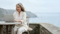 Middle-aged woman with long gray hair, Caucasian ethnicity, wearing cream linen clothing, working on laptop at outdoor stone table overlooking coastal cliffs and ocean. Remote work and digital nomad lifestyle concept.
