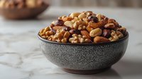 Mixed nuts and dried fruits in ceramic bowl on marble counter for healthy snacking and cooking ingredients.
