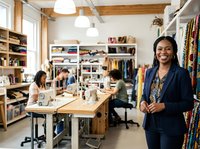 Female African American fashion designer or entrepreneur smiling in modern studio workspace with colleagues, clothing racks, and design materials for fashion business and creative industry.