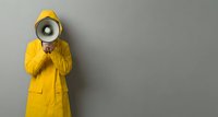 Person in bright yellow raincoat holding megaphone against gray wall background for communication and announcement concepts.